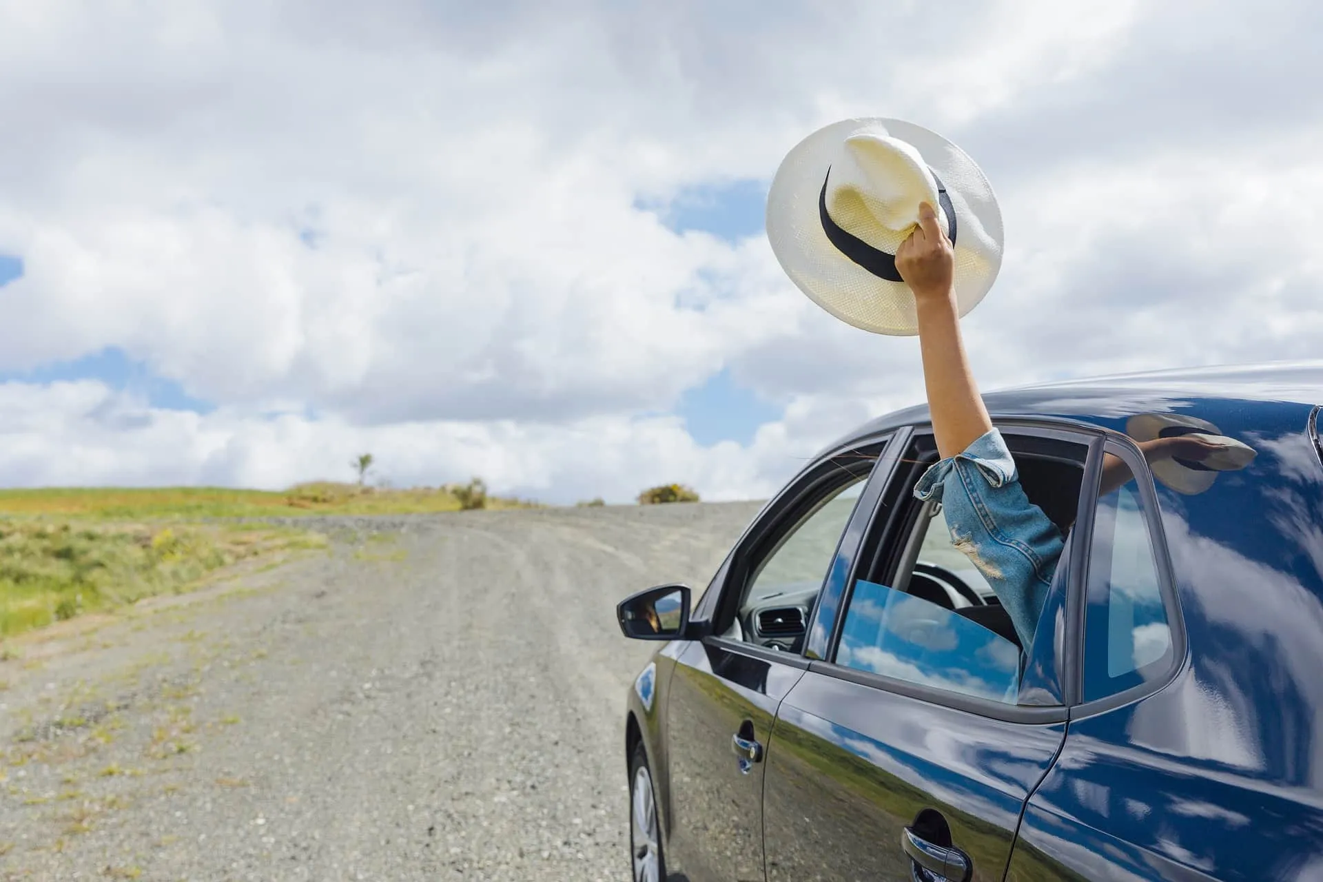 A person waving a hat from a car window while driving along a gravel road under a bright, cloudy sky.