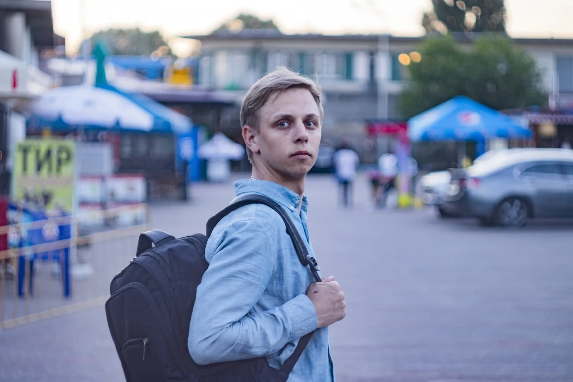 A young man with a backpack walking through a street, looking back at the camera in an outdoor urban setting.