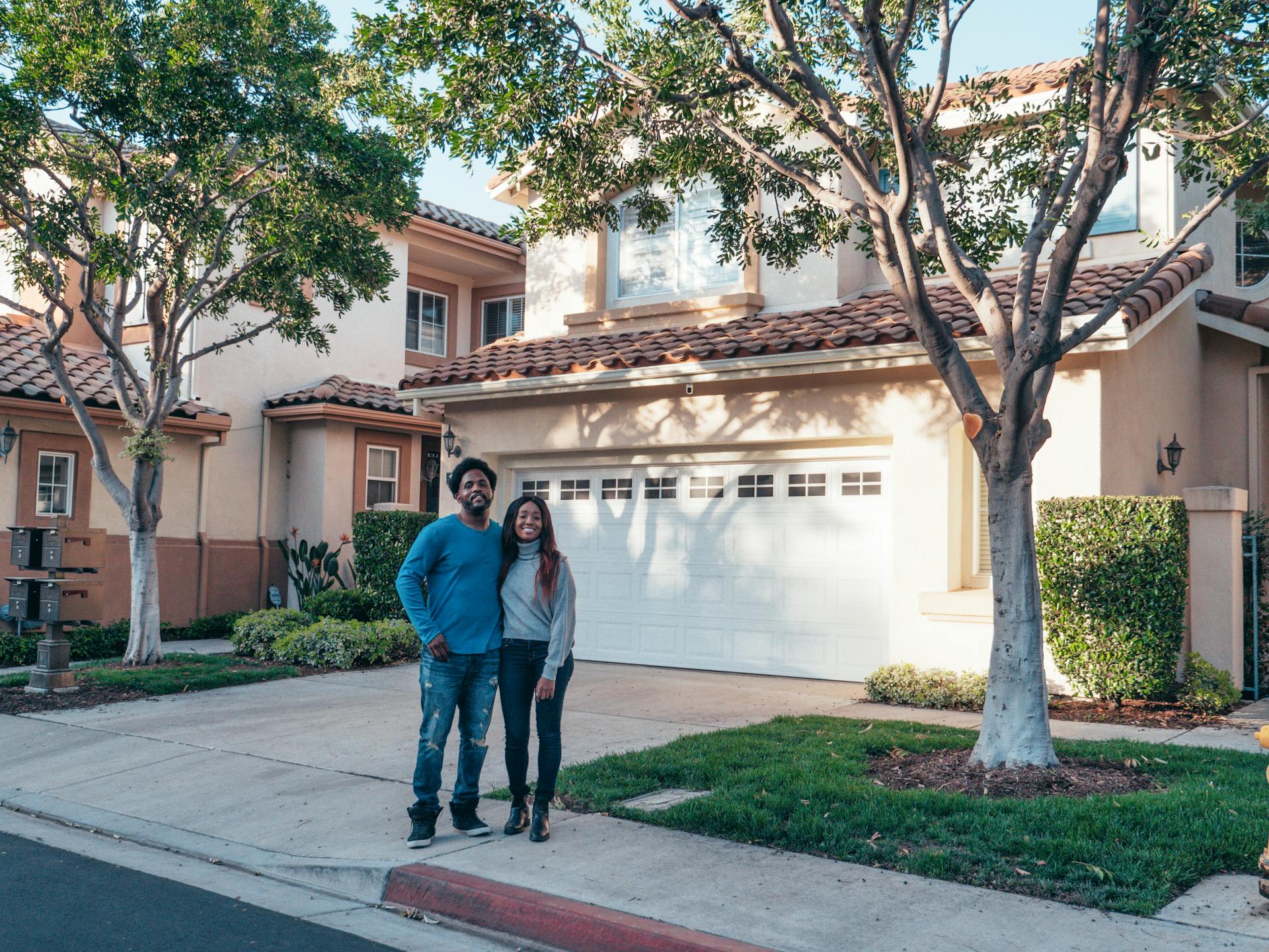 Smiling couple standing in front of their suburban home surrounded by trees, representing the comfort of suburban living.