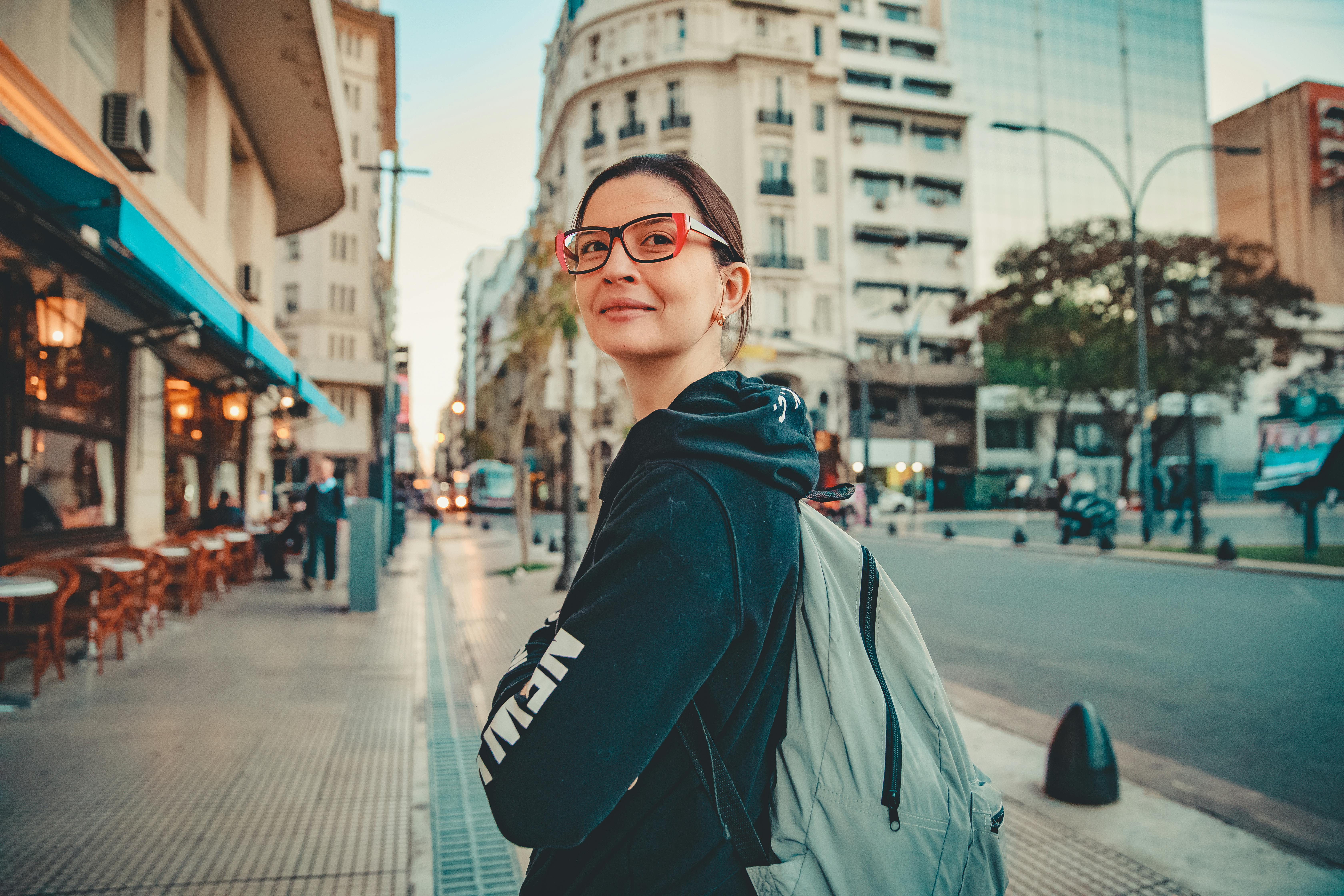 A woman wearing glasses and a backpack walks along a city street.