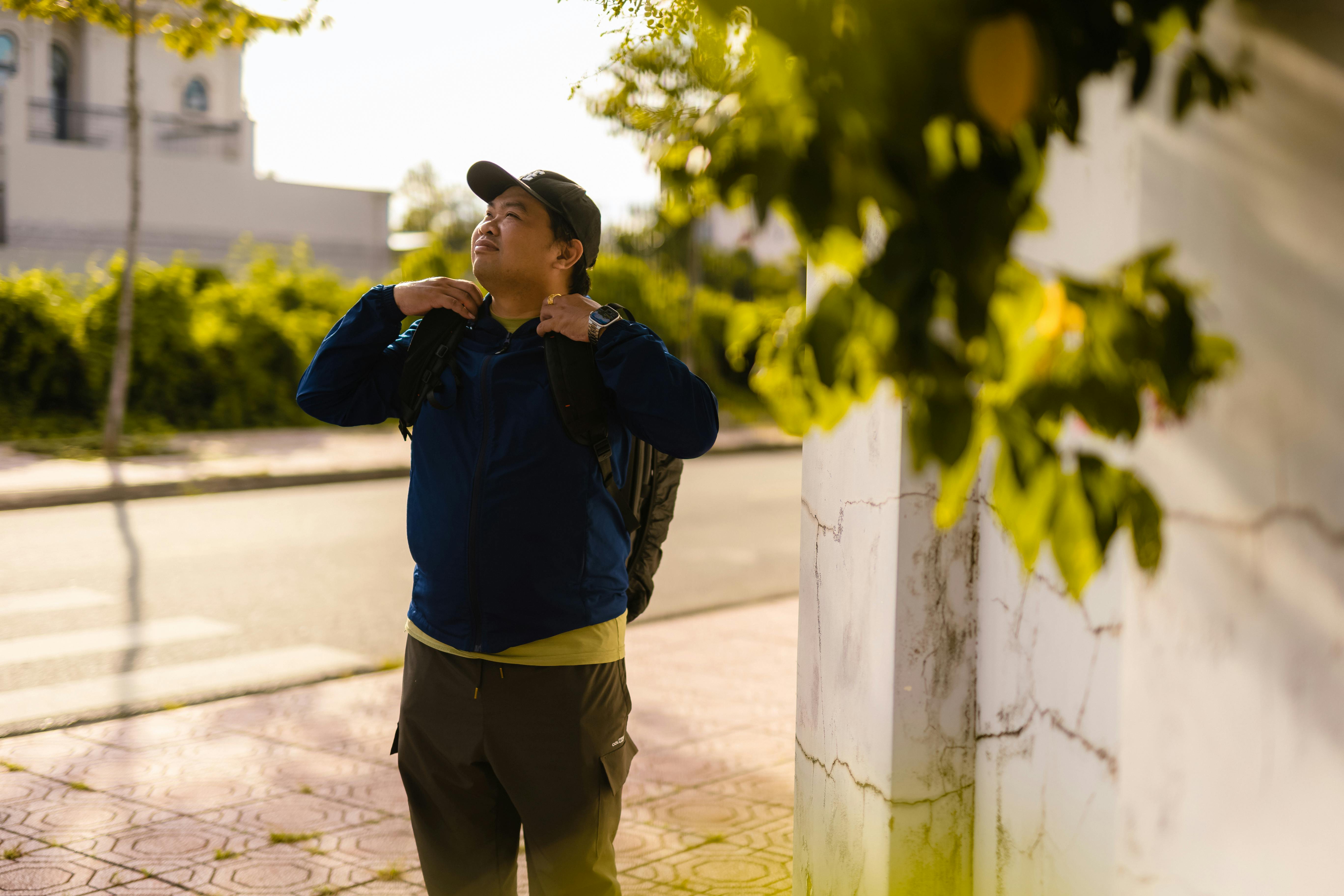 A person with a backpack and a cap stands on a sunlit street.
