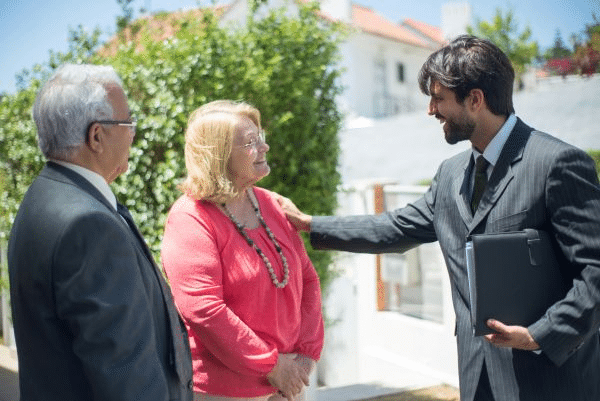 three people engaged in an outdoor conversation, with two older adults facing a younger man in a suit holding a folder.
